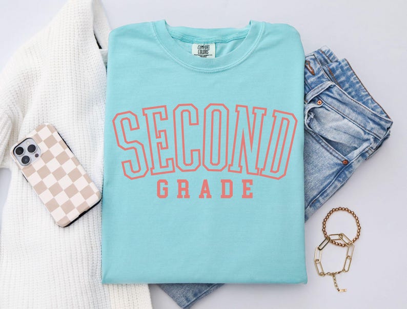 Puede incluir: Una camiseta azul claro con las palabras "SECOND GRADE" en letras color coral. La camiseta est&aacute; doblada junto a unos vaqueros azules, un su&eacute;ter blanco, una funda de tel&eacute;fono y una pulsera dorada. Marca Comfort Colors.