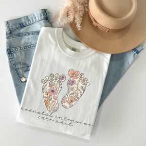 Puede incluir: Camiseta blanca con un dise&ntilde;o floral de dos huellas y el texto "neonatal intensive care unit".