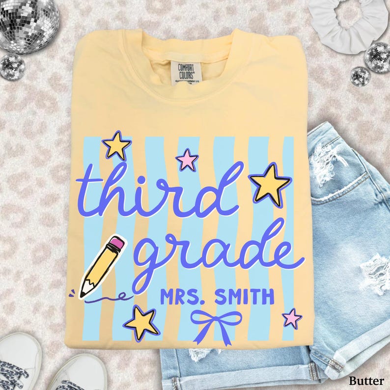May include: Butter-colored t-shirt with the words "third grade" in purple script, blue vertical stripes, and star graphics. The text "MRS. SMITH" and a bow are below the words. A pencil graphic is also included.