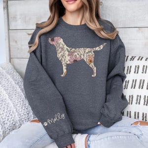 May include: A dark gray sweatshirt with a floral print labrador retriever design. The sweatshirt has a long sleeve and a crew neck. The text "Molly" is printed on the left sleeve.