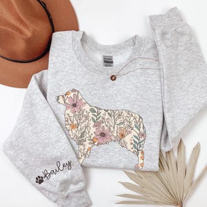 Puede incluir: Una sudadera gris con un dise&ntilde;o floral de una silueta de perro. El perro est&aacute; delineado en negro y relleno de flores rosas, amarillas y verdes. La palabra "Bailey" est&aacute; escrita en la manga con una huella de pata.