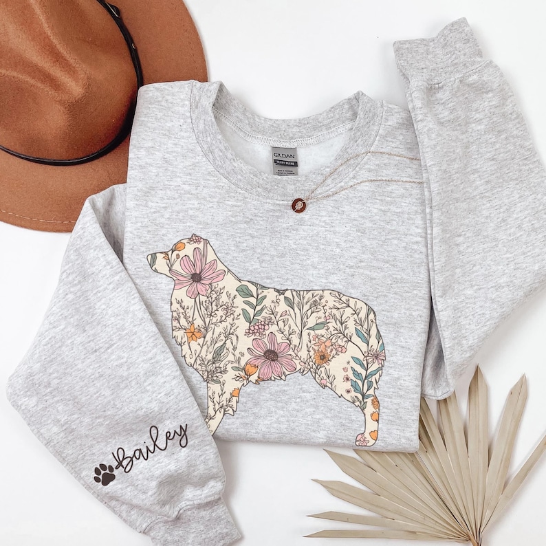 Puede incluir: Una sudadera gris con un dise&ntilde;o floral de una silueta de perro. El perro est&aacute; delineado en negro y relleno de flores rosas, amarillas y verdes. La palabra "Bailey" est&aacute; escrita en la manga con una huella de pata.