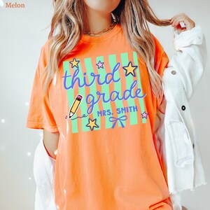 May include: An orange t-shirt with the words "third grade" in blue script, a yellow pencil, and stars. The shirt also includes the name "Mrs. Smith" and a bow. The shirt is a comfortable, casual style.