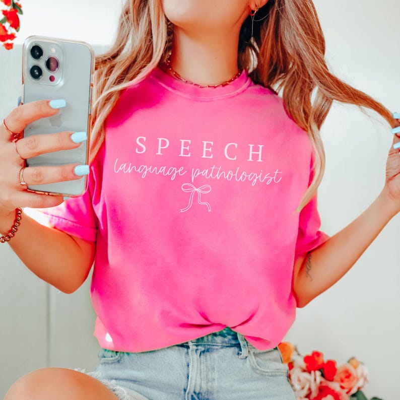 K&ouml;nnte beinhalten: Leuchtend pinkfarbenes T-Shirt mit dem wei&szlig;en Schriftzug "SPEECH language pathologist". Unter dem Text befindet sich eine wei&szlig;e Schleifen-Grafik. Das Shirt ist einfarbig und hat einen Rundhalsausschnitt.