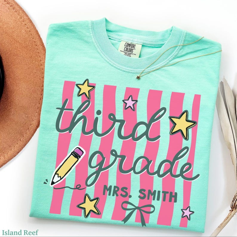 May include: A light teal Comfort Colors t-shirt with pink vertical stripes and the words "third grade" in a whimsical font. The shirt features star and pencil graphics, and the name "MRS. SMITH" below. A gold necklace with a star pendant is on the shirt.