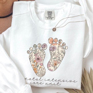 May include: White sweatshirt with a floral design of two footprints and the text "natal intensive care unit".