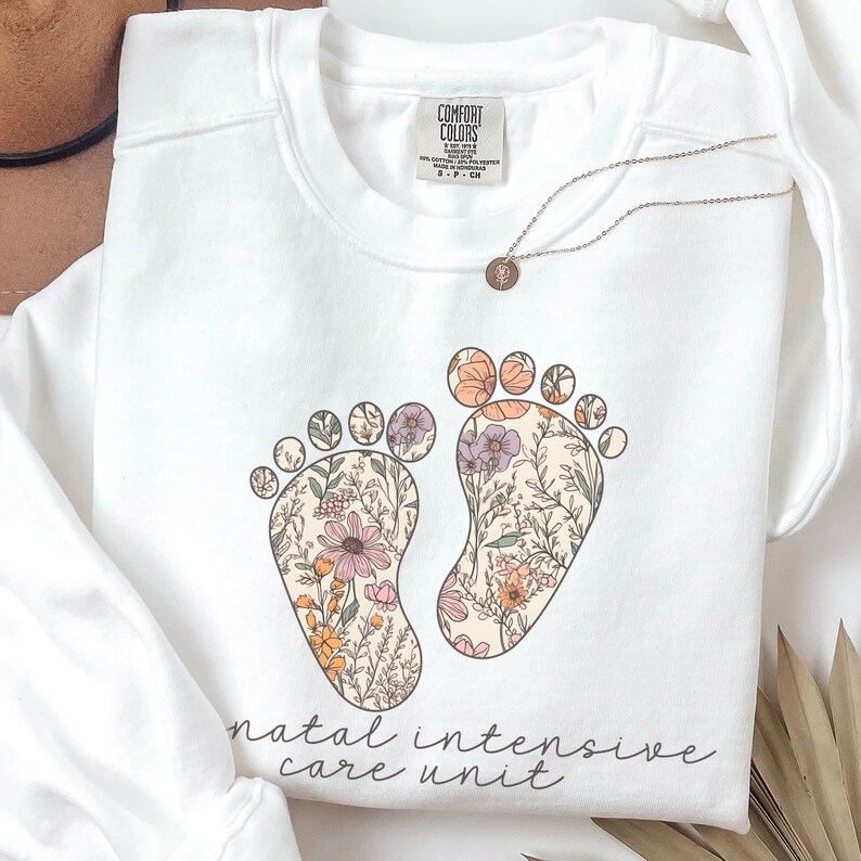 May include: White sweatshirt with a floral design of two footprints and the text "natal intensive care unit".