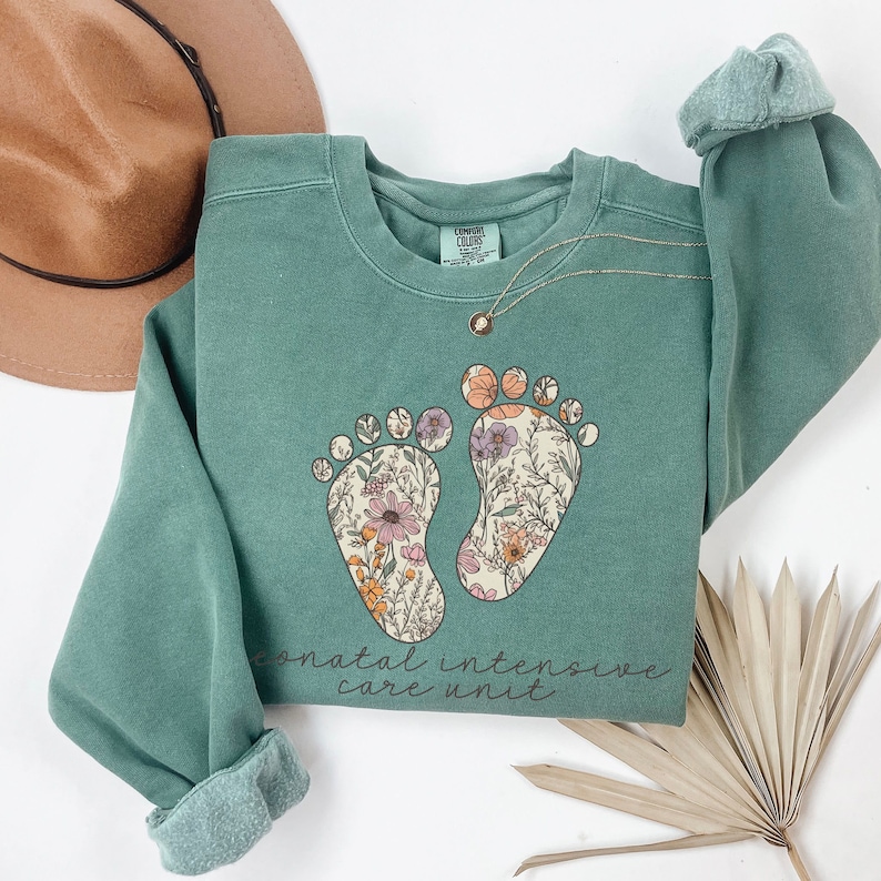 May include: A teal green sweatshirt with a floral print of two footprints and the text "neonatal intensive care unit".