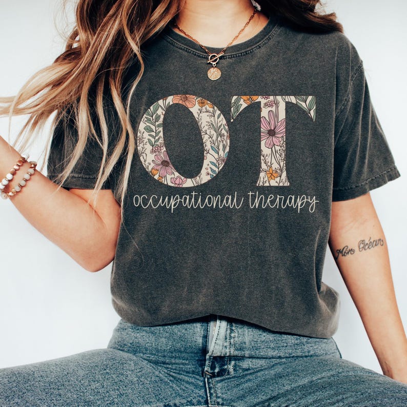 K&ouml;nnte beinhalten: Ein dunkelgraues T-Shirt mit den Buchstaben "OT" in Wei&szlig; mit einem floralen Muster innerhalb der Buchstaben. Der Text "occupational therapy" steht unter den Buchstaben.