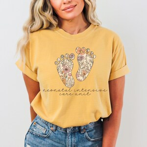 Puede incluir: Una camiseta amarilla con un dise&ntilde;o floral de dos huellas y el texto "neonatal intensive care unit".
