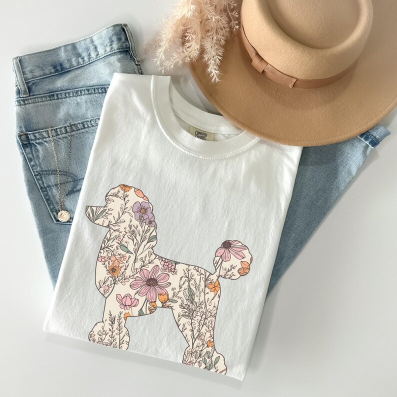 May include: White t-shirt with a floral poodle design. The poodle is outlined in black and filled with pink, orange, and purple flowers.