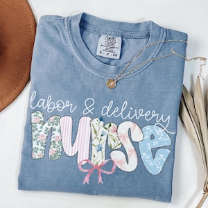 May include: A light blue t-shirt with the text "labor & delivery nurse" in white lettering. The word "nurse" is made up of colorful patchwork letters with floral and ribbon designs.