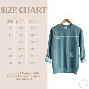 May include: Size chart for a unisex garment-dyed sweatshirt in sizes S through 3XL. The chart shows the width and length measurements in inches for each size. The sweatshirt is a light blue color.