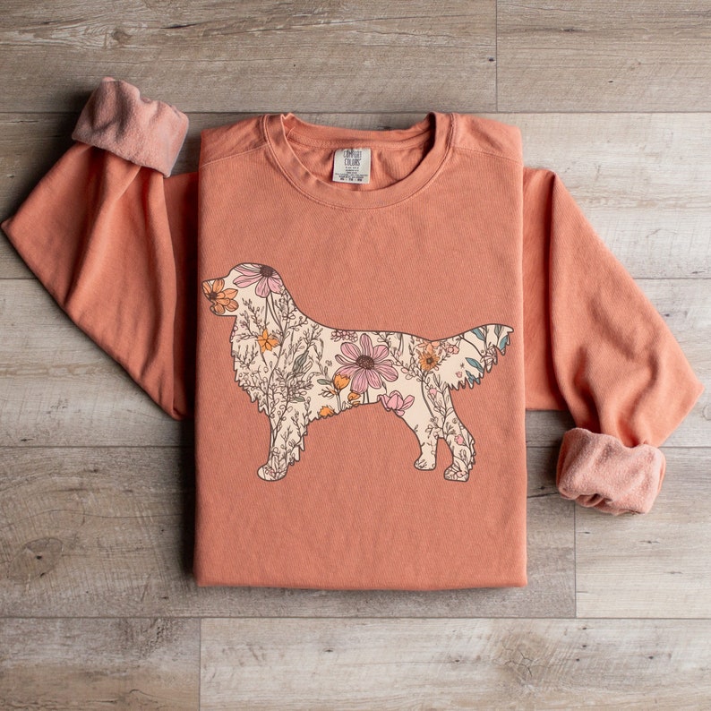 May include: A peach-colored sweatshirt with a floral design of a golden retriever. The floral design is in shades of pink, yellow, and green.