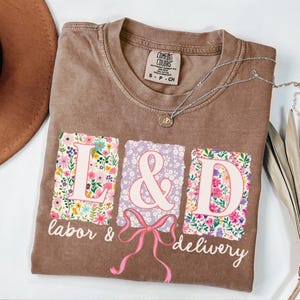 May include: A brown t-shirt with a floral print design that spells out "L&D" with the words "labor & delivery" underneath.