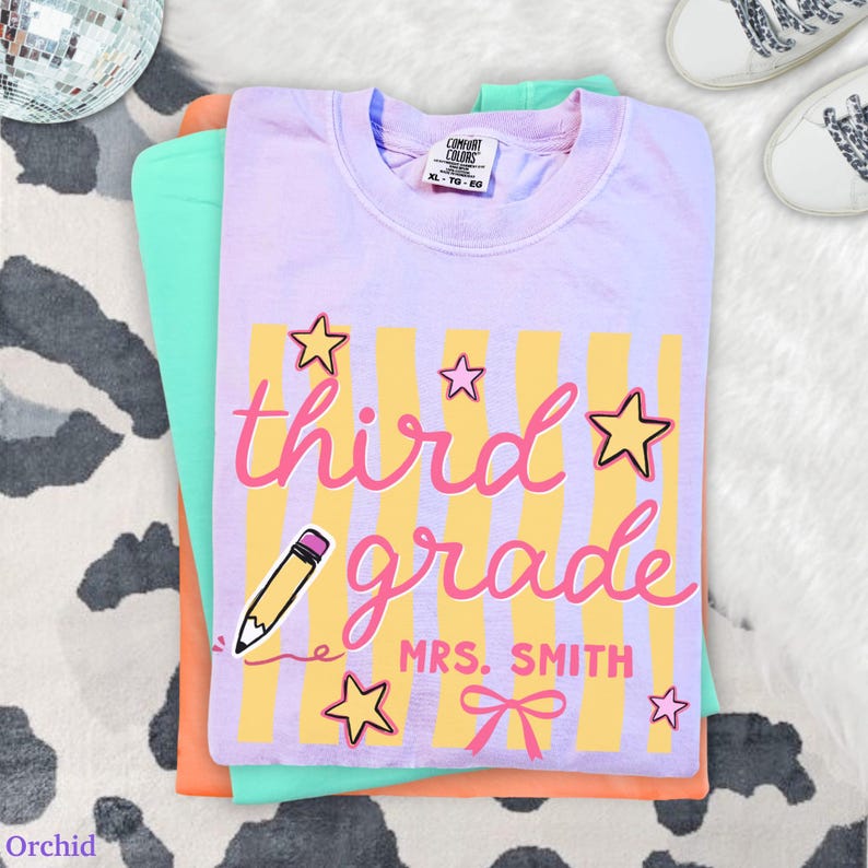 May include: A stack of three t-shirts in shades of orchid, orange, and mint green. The top shirt is orchid and features the words "third grade" in pink script with yellow stripes and star accents. The text "MRS. SMITH" is also visible.
