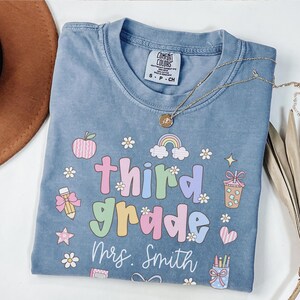 Op de afbeelding: Een blauw Comfort Colors T-shirt met de woorden "third grade" in kleurrijke letters, samen met een regenboog, een appel en andere schoolthema-afbeeldingen. De naam "Mrs. Smith" staat eronder gedrukt.