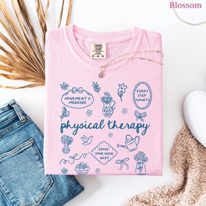 Puede incluir: Una camiseta rosa claro con las palabras "physical therapy" y varias ilustraciones azules. La camiseta tambi&eacute;n presenta las frases "Movement is Medicine" y "Every Step Counts". Un delicado collar descansa sobre la camiseta, junto a unos vaqueros azules.