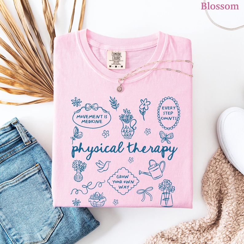 Puede incluir: Una camiseta rosa claro con las palabras "physical therapy" y varias ilustraciones azules. La camiseta tambi&eacute;n presenta las frases "Movement is Medicine" y "Every Step Counts". Un delicado collar descansa sobre la camiseta, junto a unos vaqueros azules.