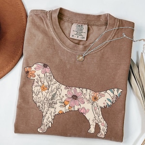May include: A brown t-shirt with a floral design of a golden retriever. The shirt has the text "COMFORT COLORS" and "S - P - CH" on the tag.