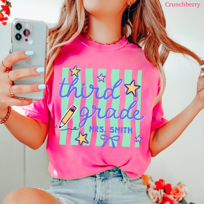 May include: A bright pink t-shirt with the words "third grade" in a whimsical font, accented with stars, a pencil, and green stripes. The name "MRS. SMITH" is below the words. A fun back-to-school design.