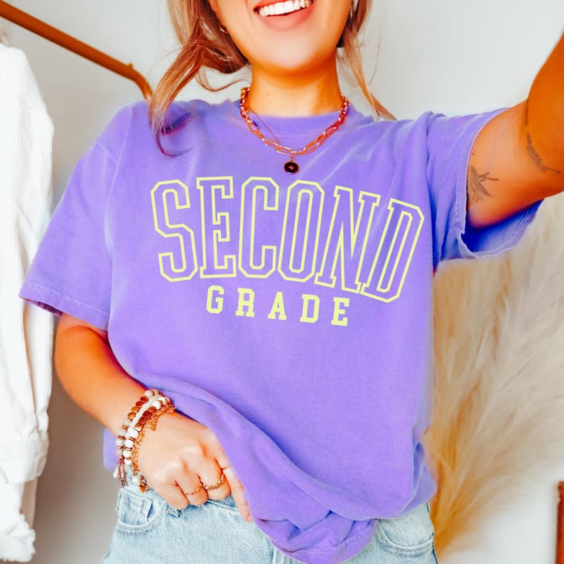 Puede incluir: Una camiseta morada con las palabras "SECOND GRADE" en letras amarillas. El texto est&aacute; delineado en amarillo m&aacute;s oscuro. La camiseta tiene cuello redondo y es de estilo casual. La camiseta es de color s&oacute;lido.
