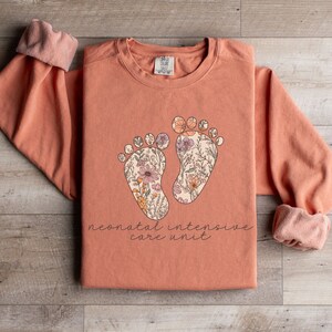 May include: A peach-colored long-sleeved sweatshirt with a floral design of two baby footprints and the text "neonatal intensive care unit".