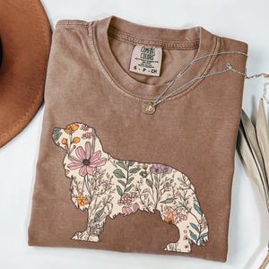 Può includere: Una maglietta marrone con un design floreale a forma di Cavalier King Charles Spaniel. La maglietta ha il testo "COMFORT COLORS" sull'etichetta.