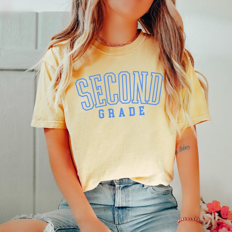 Puede incluir: Camiseta amarilla de manga corta con las palabras "SECOND GRADE" impresas en azul. La camiseta tiene un aspecto ligeramente descolorido. El texto est&aacute; en negrita, en una fuente de estilo universitario.