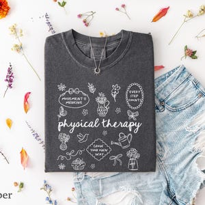 Puede incluir: Una camiseta gris oscuro con las palabras "physical therapy" y garabatos blancos. La camiseta est&aacute; tendida con unos pantalones cortos de mezclilla y un collar plateado. Flores y hojas de colores esparcidas.