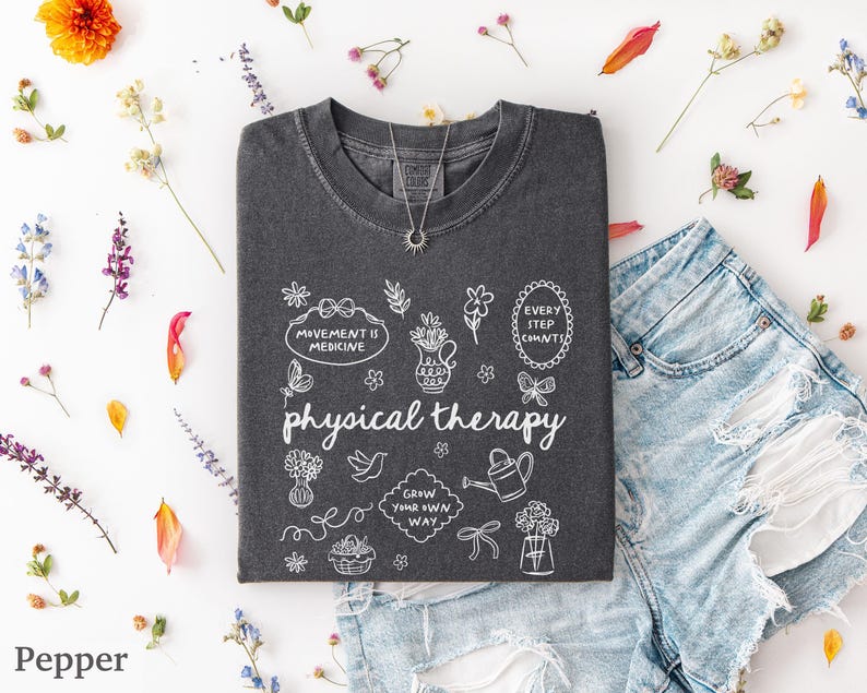 Puede incluir: Una camiseta gris oscuro con las palabras "physical therapy" y garabatos blancos. La camiseta est&aacute; tendida con unos pantalones cortos de mezclilla y un collar plateado. Flores y hojas de colores esparcidas.
