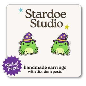 May include: A pair of handmade earrings featuring green cartoon frogs wearing purple wizard hats with gold trim and stars. The earrings are nickel-free with titanium posts. The text "Stardoe Studio" is at the top.