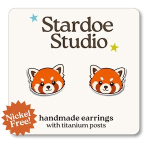 May include: A pair of stud earrings featuring red panda faces with orange, white, and brown details. The earrings are displayed on a white card labeled Stardoe Studio with a star graphic and text stating handmade earrings with titanium posts and Nickel Free.