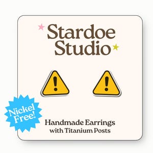 May include: A pair of handmade earrings with titanium posts, featuring a yellow triangle warning sign with a black exclamation point. The earrings are displayed on a white card with the text "Stardoe Studio" and "Nickel Free!"