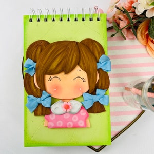 May include: A green spiral notebook with a 3D felt illustration of a girl with brown hair, blue bows, and a pink dress with white polka dots.