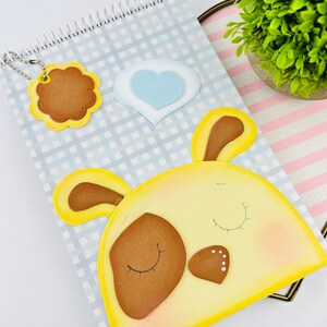 May include: A blue and white checkered notebook with a yellow and brown felt animal applique. The animal has brown eyes and a brown nose. The notebook has a spiral binding and a small felt flower with a key ring attached.