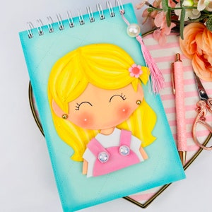 May include: A turquoise blue spiral notebook with a 3D girl character on the front. The girl has blonde hair, a pink dress, and a pink flower in her hair. The notebook has a white tassel.