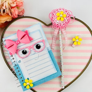 May include: A blue and white spiral notebook with a pink bow, pink glasses, and a white face with black eyes. The notebook is on a pink and white striped heart-shaped surface. A pink pencil with a yellow flower on top is next to the notebook.