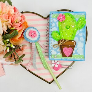 May include: A heart-shaped notebook with a blue and white cover featuring a green cactus with a pink flower and a pink heart. The notebook is decorated with white polka dots and a light green ribbon. A pink flower with a white center is attached to a light green and silver ribbon.