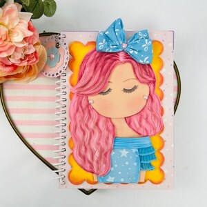 May include: A pink and blue spiral notebook with a 3D paper doll on the cover. The doll has pink hair, blue eyes, and is wearing a blue dress with white stars. The doll is wearing a blue bow with a white center.