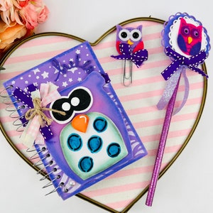 May include: A purple owl-themed notebook with a spiral binding, a purple pencil with glitter, and two purple owl-shaped paper clips. The notebook has a white background with stars and the word "little" written in purple.