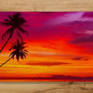 Könnte beinhalten: Silhouette von zwei Palmen vor einem leuchtenden Sonnenuntergang mit Orange-, Rosa- und Lilatönen. Der Horizont ist mit einem ruhigen Gewässer im Vordergrund sichtbar.