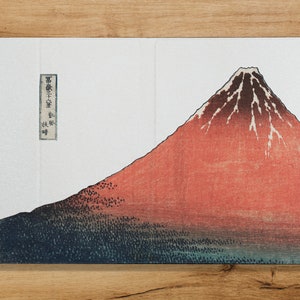 Puede incluir: Una funda de teléfono blanca con una imagen roja y negra del Monte Fuji, una famosa montaña japonesa. La funda tiene una pequeña etiqueta de texto japonés en el lado izquierdo.