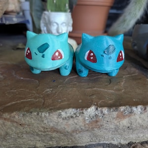 Puede incluir: Dos macetas Bulbasaur de color verde azulado. Cada maceta presenta detalles en rojo y blanco en la cara y una marca verde oscuro en la parte posterior. Las macetas están sobre una superficie de piedra, con plantas y macetas en el fondo.