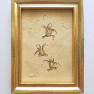 Puede incluir: Una obra de arte enmarcada que presenta tres crías de tortuga marina nadando en un entorno arenoso. Las tortugas están representadas en tonos marrones y beige, con un patrón de caparazón detallado. El marco es dorado.