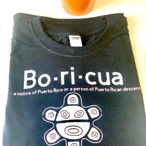 May include: Black t-shirt with white text that reads "Bo-ri-cua: a native of Puerto Rico or a person of Puerto Rican descent". A white stylized image of a Puerto Rican flag symbol is below the text.