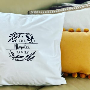 May include: White square pillow with a black floral design and the text "The Morales Family". A yellow pillow with pom-poms and a white pillow with a beige trim are also visible. Home decor.