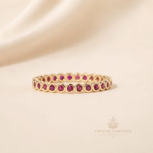 Op de afbeelding: Een gouden eeuwigheidsring met een doorlopende rij ronde, levendige roze robijnen in individuele ronde zettingen. De ring heeft een gepolijste metalen afwerking en een uniform ontwerp, met de tekst Crystal Carvings India zichtbaar in de hoek.