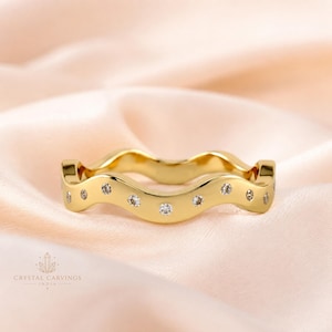 Può includere: Un anello in oro con una silhouette ondulata continua. La superficie dell'anello è lucida e incastonata con una serie di piccole pietre rotonde e trasparenti, distanziate uniformemente lungo il design sinuoso. Il testo Crystal Carvings India è visibile.