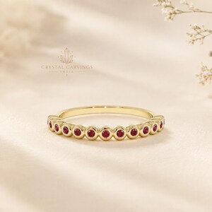 May include: A gold band ring featuring a series of small, round, bezel-set red rubies arranged in a continuous row along the top half. The metal has a polished finish, and the text CRYSTAL CARVINGS INDIA is visible above the ring.